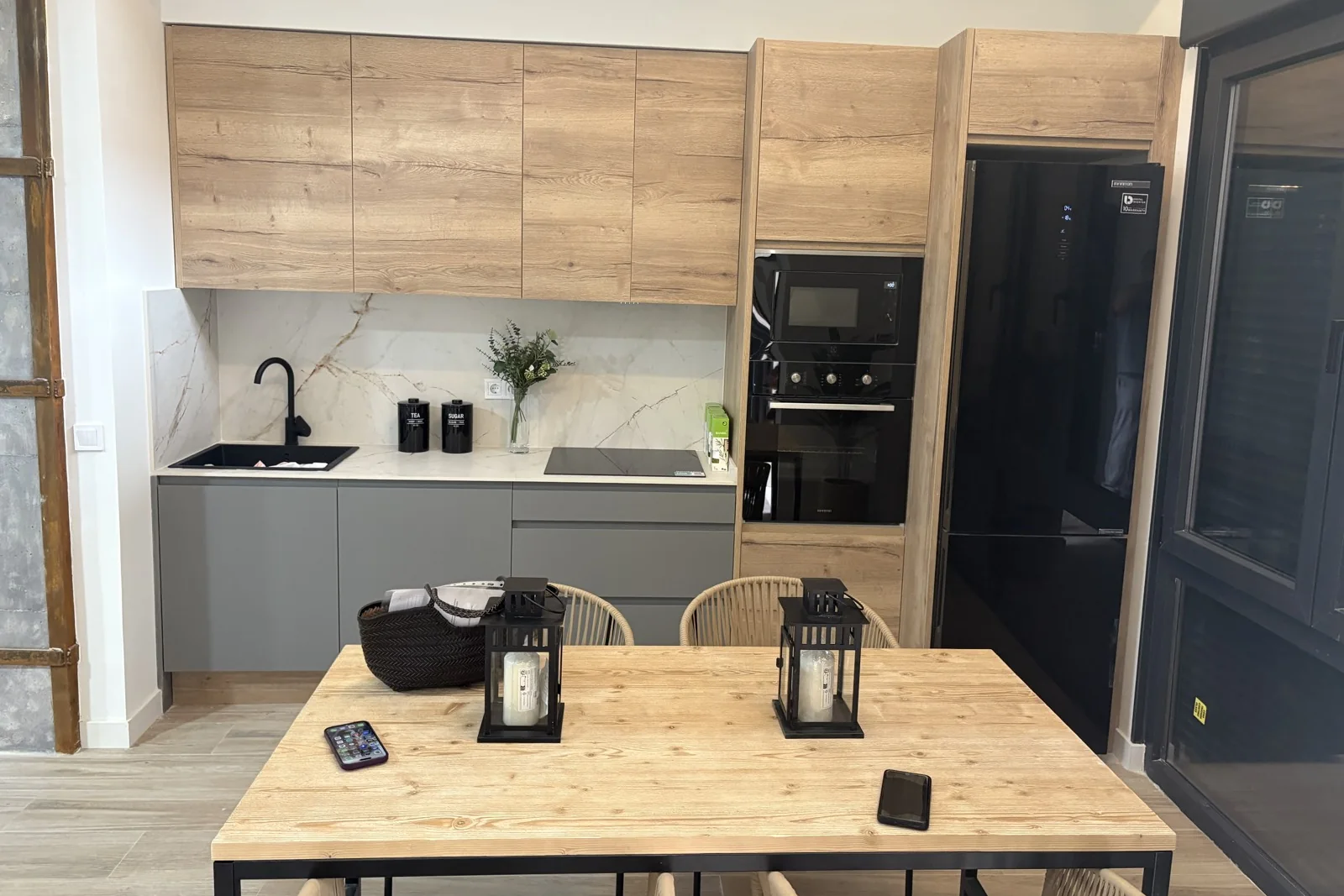 Cocina abierta al comedor en un local convertido en vivienda: muebles altos de madera, columna de horno, frigorífico negro y mesa de madera