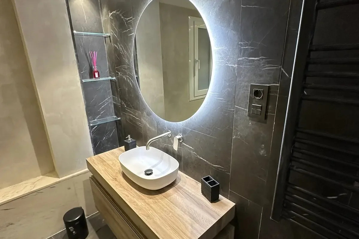 Mueble de lavabo y espejo iluminado. Lavabo sobre mueble volado en tono madera, espejo redondo iluminado, toallero negro y revestimiento gris oscuro veteado.