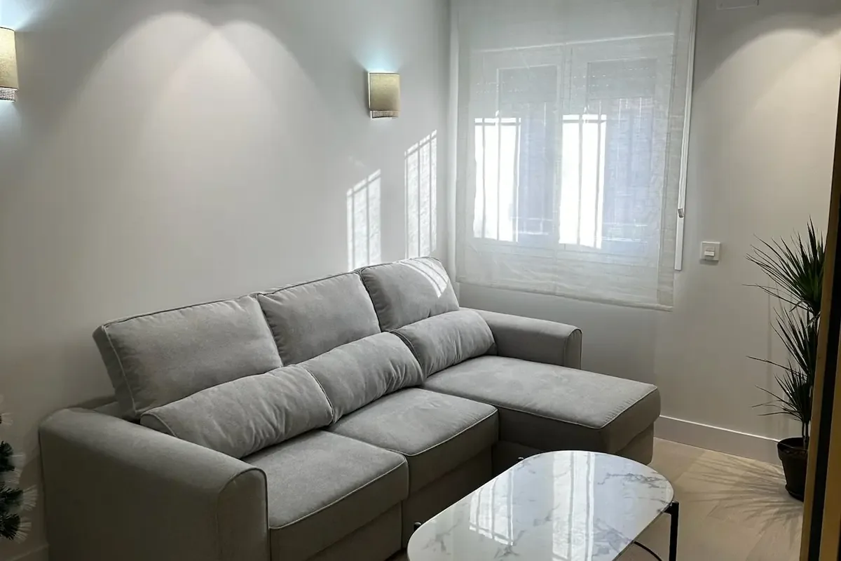 Salón con sofá rinconera y apliques. Salón con sofá gris claro, mesa de centro de tapa veteada, apliques de pared y suelo claro en lamas.