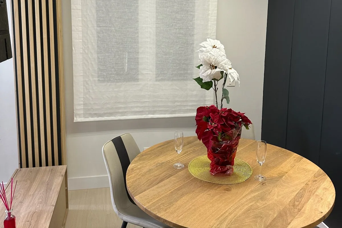 Comedor reformado con mesa redonda de madera junto a la ventana, pilar forrado de listones y línea de luz en el techo