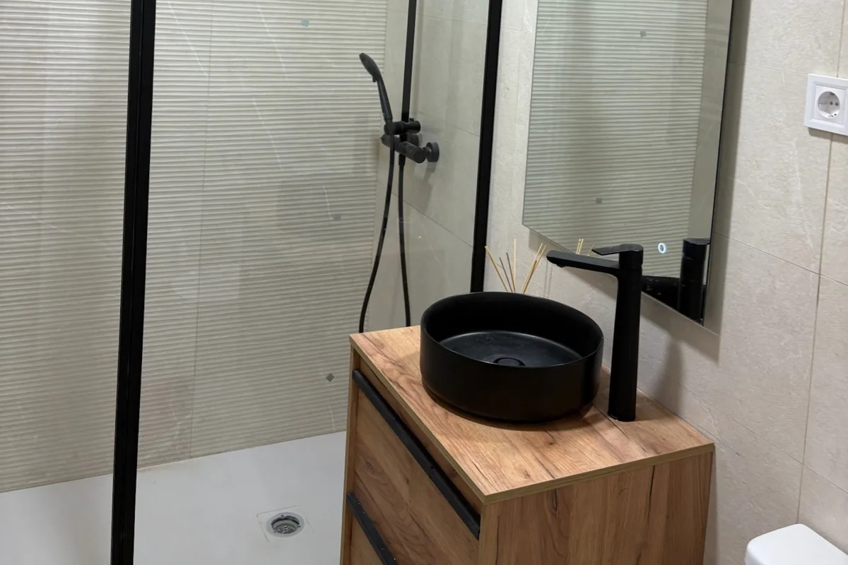 Baño con ducha a ras de suelo, mampara de cristal con perfil negro, columna de ducha negra y lavabo redondo negro sobre mueble de madera