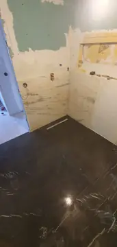 Colocación del revestimiento de la ducha. Piezas claras con veta gris en la zona de ducha, con canaleta de desagüe y pavimento oscuro pulido ya colocado.