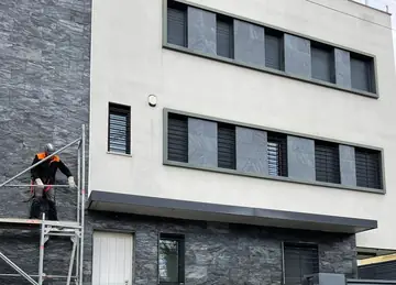 Fachada con andamio durante el aplacado. Aplacado de piedra oscura de cara partida en planta baja, con un operario en el andamio y paneles grises entre las ventanas.