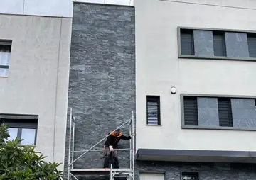 Andamio en la franja aplacada. Franja vertical de piedra oscura de cara partida entre dos fachadas claras, con un operario trabajando desde el andamio.
