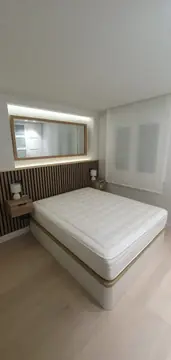 Dormitorio doble con espejo iluminado. Cama doble ante panel de listones en tono madera, mesillas voladas y espejo enmarcado en hornacina con luz indirecta.