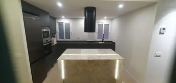 Isla de cocina veteada con luz en las aristas. Isla con encimera y frentes claros veteados y luz integrada en las aristas, ante muebles oscuros y campana cilíndrica negra.