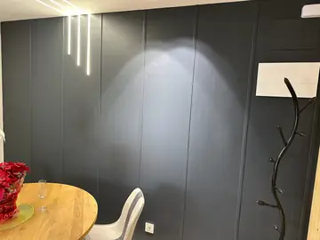 Pared de paneles oscuros con luz lineal. Revestimiento de paneles oscuros con juntas verticales y tres líneas de luz empotradas, junto a la mesa del comedor.