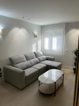 Salón con sofá rinconera y apliques. Salón con sofá gris claro, mesa de centro de tapa veteada, apliques de pared y suelo claro en lamas.