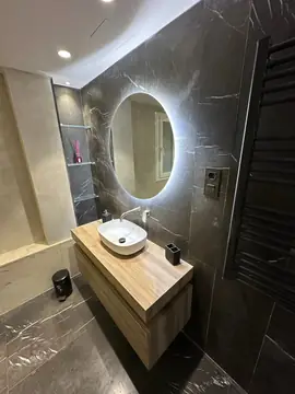 Mueble de lavabo y espejo iluminado. Lavabo sobre mueble volado en tono madera, espejo redondo iluminado, toallero negro y revestimiento gris oscuro veteado.
