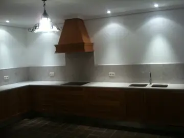 Cocina en L con encimera y frontal claros. Encimera clara y frontal de grano gris a media altura en una cocina con muebles en tono madera.