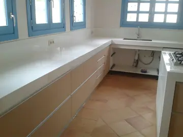 Encimera blanca en L bajo las ventanas. Encimera blanca pulida en L con fregadero bajo encimera, con el mueble del seno aún sin puertas.