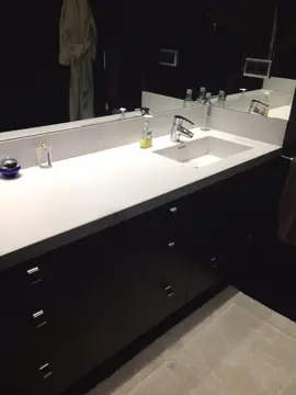 Encimera de baño blanca con lavabo integrado. Encimera blanca con lavabo integrado y copete sobre mueble oscuro de cajones, bajo espejo.