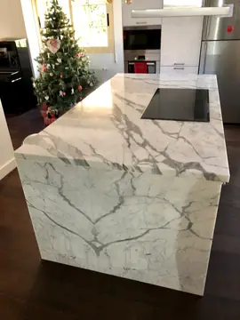 Isla blanca veteada con costado en cascada. Isla de cocina con encimera y costado en cascada blancos con veta gris marcada, con la placa encastrada.