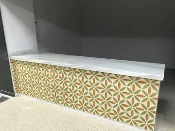 Mostrador visto en escorzo. Mostrador con encimera blanca de veta gris y frente de baldosa decorada, sobre pavimento claro.