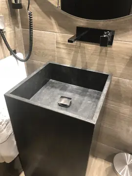 Lavabo exento negro con grifería mural. Lavabo cuadrado negro de acabado mate con grifo empotrado en la pared y revestimiento en tono madera.