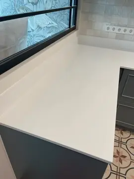 Encimera blanca con copete bajo la ventana. Tramo de encimera blanca lisa con copete del mismo material junto a una ventana interior con vidrio decorado.