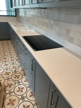 Tramo de encimera blanca con placa. Encimera blanca lisa con placa negra encastrada y fregadero al fondo, frente a un azulejo claro de aspecto artesanal.