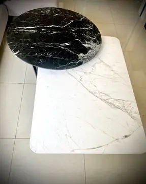 Mesas de centro vistas desde arriba. Tapa circular negra con vetas blancas sobre tapa rectangular blanca con vetas grises, en un salón de pavimento claro.
