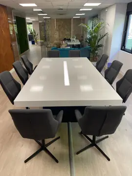 Sala de reuniones con mesa blanca. Mesa de reuniones con tapa blanca brillante y franja de luz central, rodeada de sillas tapizadas en gris.