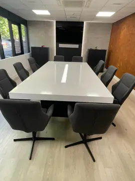 Sala de reuniones con mesa de canto grueso. Mesa de tapa blanca brillante y canto grueso con franja de luz central, rodeada de sillas en una sala luminosa.
