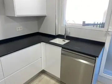 Cocina blanca con encimera negra texturizada. Encimera negra de acabado texturizado en L, con fregadero blanco bajo encimera y escurridor ranurado.