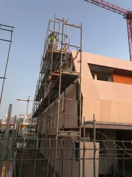 Fachada aplacada con andamio y grúa. Volumen de fachada con aplacado en tono claro y andamio en el lateral, con la grúa al fondo.