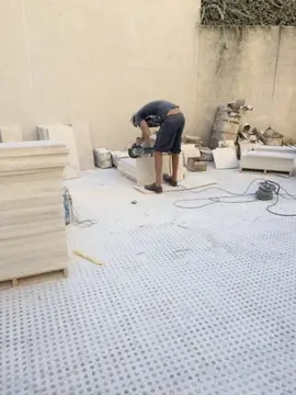 Corte de piezas a pie de obra. Operario cortando piezas en tono crema con radial, junto a palés apilados en la zona de acopio.