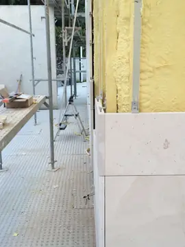 Primeras hiladas junto al andamio. Piezas en tono crema colocadas sobre el aislamiento proyectado, con andamio y mesa de trabajo a un lado.