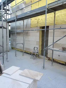 Fachada con andamio y piezas acopiadas. Paño de fachada con el aplacado avanzado alrededor de la puerta, andamio montado y piezas en primer plano.