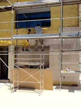 Operarios en el andamio. Dos operarios colocando piezas desde el andamio bajo una ventana, con el aplacado avanzando sobre el aislamiento.