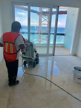 Pulido del pavimento en una habitación. Operario de espaldas pule el pavimento en tono crema con vetas, frente a un ventanal con vistas al mar y la grúa de obra.