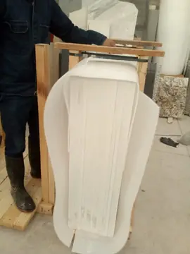 Caja de piezas blancas embaladas. Caja de madera con piezas blancas colocadas de canto y protegidas con espuma, en una nave con más tableros al fondo.