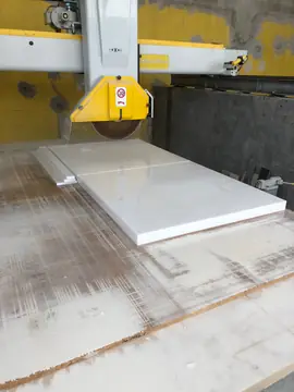 Corte de piezas blancas en taller. Cortadora de puente con el disco sobre piezas blancas pulidas recién cortadas y listones del mismo material a un lado.