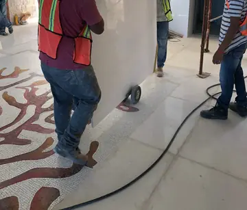 Operarios moviendo un tablero de gran formato. Cuadrilla trasladando un tablero claro sobre carro por un pavimento con mosaico de ramas rojizas.