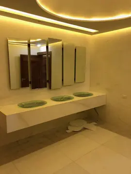 Aseo con encimera volada y espejos inclinados. Encimera blanca volada con tres lavabos de vidrio, paredes aplacadas claras y luz indirecta en el techo.