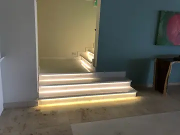 Escalera con peldaños retroiluminados. Escalera de peldaños claros con luz bajo cada huella, entre una pared azul y otra blanca.