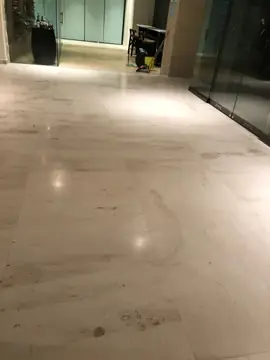 Pavimento claro con vetas en zona de paso. Pavimento claro con vetas en piezas grandes junto a cerramientos de vidrio, con útiles de limpieza al fondo.