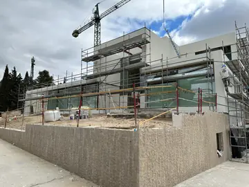 Muro de piedra y fachada con andamio. Muro exterior aplacado en piedra beige de cara labrada, con la vivienda aún con andamio y grúa.