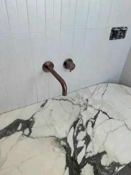 Encimera blanca de veta oscura. Encimera blanca con veta gris oscura y extremo redondeado, con grifería de pared en tono bronce.