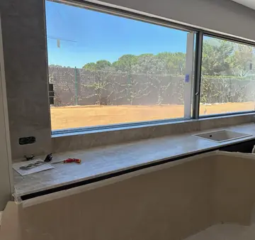 Encimera corrida bajo ventanal. Encimera y revestimiento del hueco del ventanal en gris claro veteado, con fregadero blanco y los muebles aún protegidos.