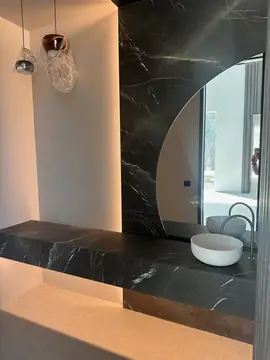 Encimera negra y espejo en media luna. Encimera volada negra con veta blanca, lavabo de apoyo blanco y espejo curvo encajado en el revestimiento.