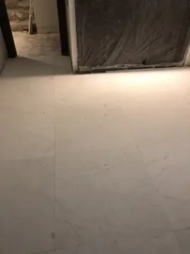 Pavimento de piezas blancas en una estancia. Suelo de piezas blancas grandes con veta gris suave, con un hueco cubierto con plástico oscuro al fondo.