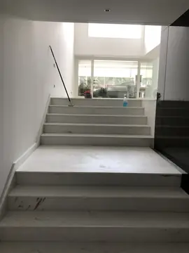 Escalera blanca hacia el salón. Tramos de peldaños blancos con veta gris y rellano en piezas grandes, con el salón al fondo durante la limpieza final.