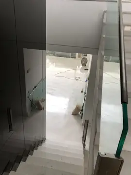 Escalera vista desde arriba en obra. Bajada de peldaños blancos entre vidrio y pared, con el salón al fondo aún en obra.