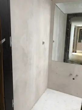 Baño en obra con espejo. Paredes en piezas grandes gris claro, espejo sobre la zona del lavabo y suelo blanco, antes de colocar sanitarios.