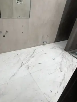 Suelo de baño blanco con veta marcada. Pavimento en piezas grandes blancas con veta gris y parda junto a una pared gris claro con tomas de agua a la vista.