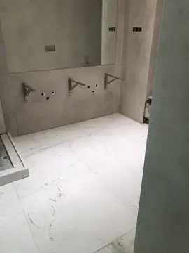 Baño en obra antes del lavabo. Pared gris claro con soportes para la encimera y suelo blanco veteado junto al plato de ducha, antes del montaje final.