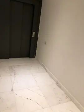 Suelo veteado junto a la puerta del ascensor. Rincón con pavimento blanco de veta gris marcada y rodapié a juego frente a la puerta oscura del ascensor.
