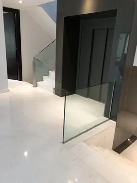Distribuidor con escalera y vidrio. Pavimento blanco pulido junto al hueco de la escalera, con barandilla de vidrio y núcleo de ascensor oscuro.