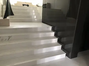 Escalera blanca junto a vidrio oscuro. Peldaños blancos con veta gris que suben entre una escultura y un paño de vidrio oscuro.